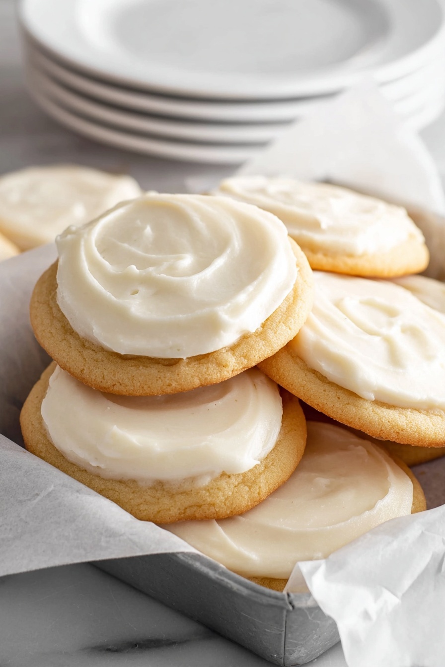 The image shows a stack of round cookies with smooth, creamy white frosting spread generously on top of each one, covering the entire top surface. The cookies have a golden-brown color and slightly soft texture, piled together inside a white paper-lined gray tray. In the background, there are several clean white plates stacked on a white marbled surface. photo taken with an iphone --ar 2:3 --v 7