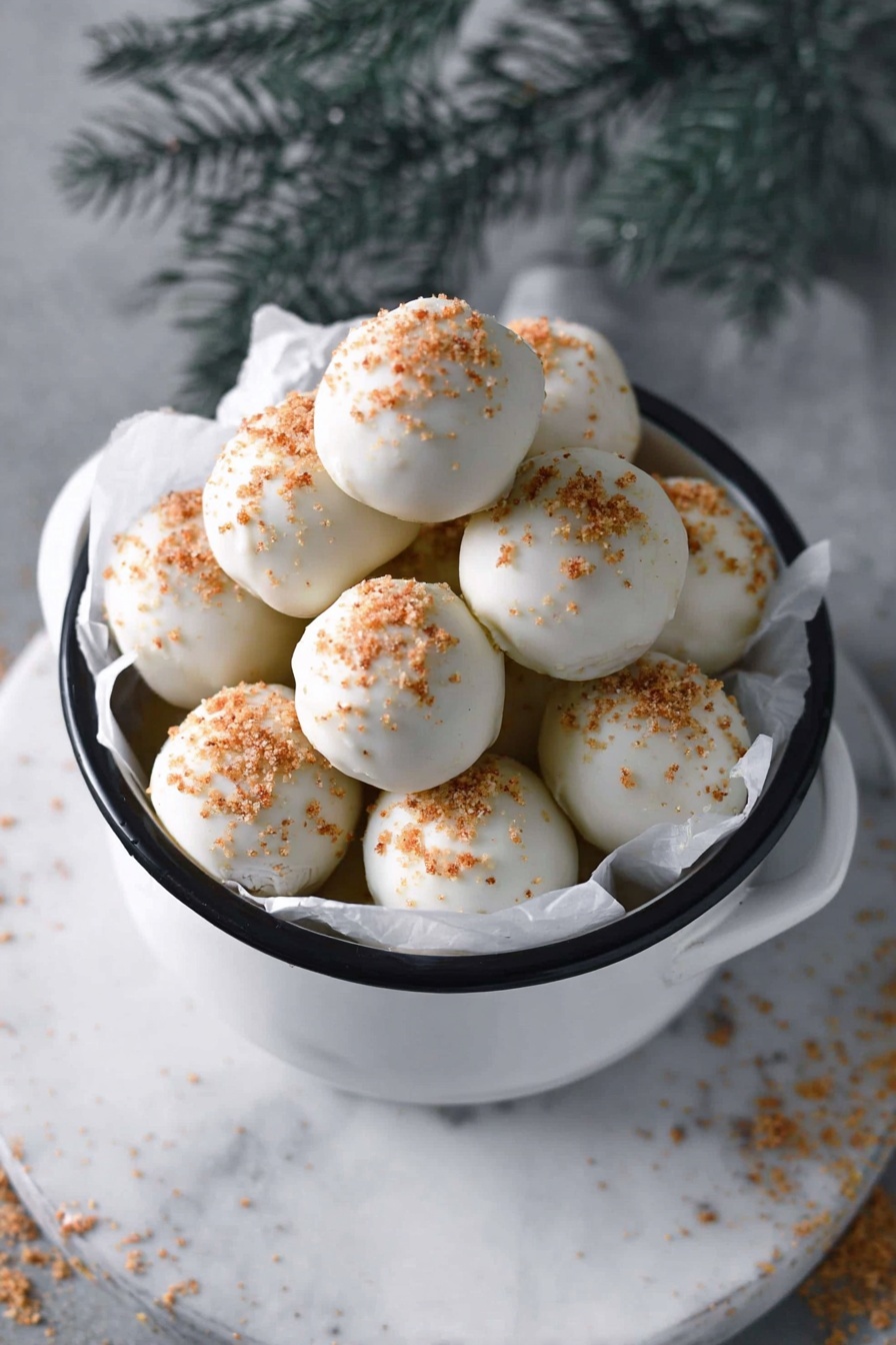 A white bowl filled with round white-coated balls stacked in a pyramid shape, each ball sprinkled with small brown crumbs on top, sitting on a white marbled surface. The bowl has a black handle and is lined with white parchment paper, and near the bowl, there are two stacked rectangular brown cookies with patterns and the word